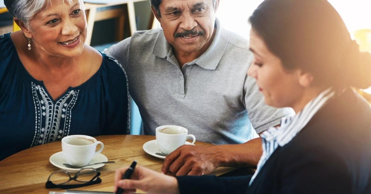 An older couple listens to their female advisor explain financial documents at a small meeting table.