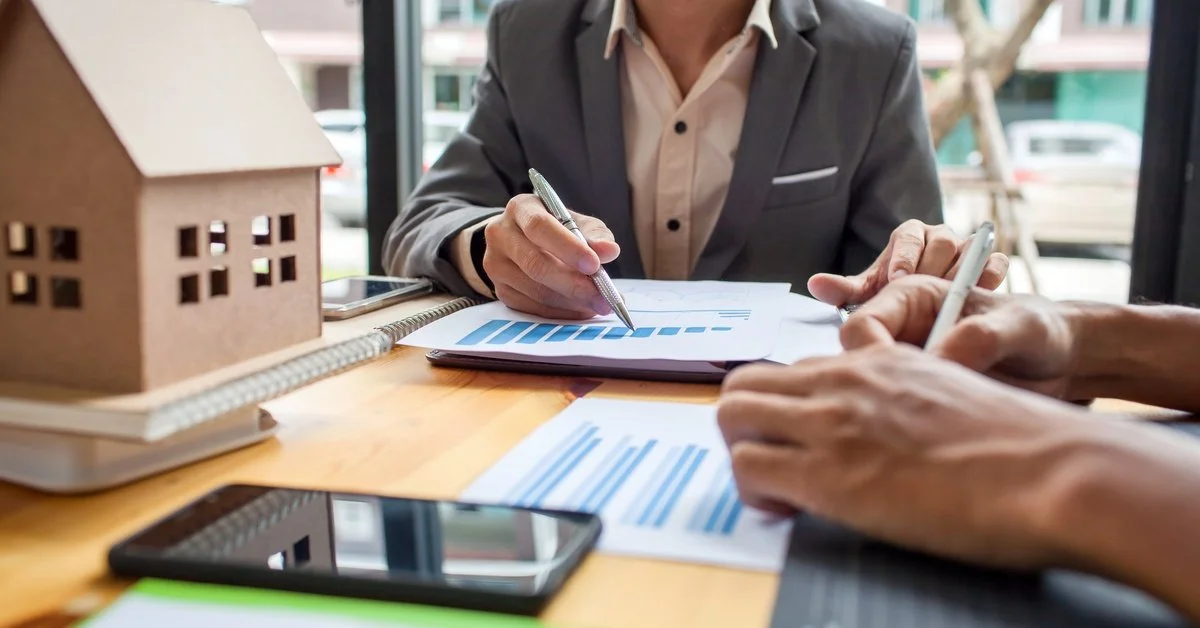 A focus shot of a desk scattered with documents, a phone, and a model house; two people are meeting at the desk.