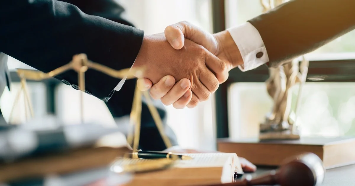 Two professionals shake hands over a desk with a gavel and scales of justice, marking a legal agreement in an office setting.
