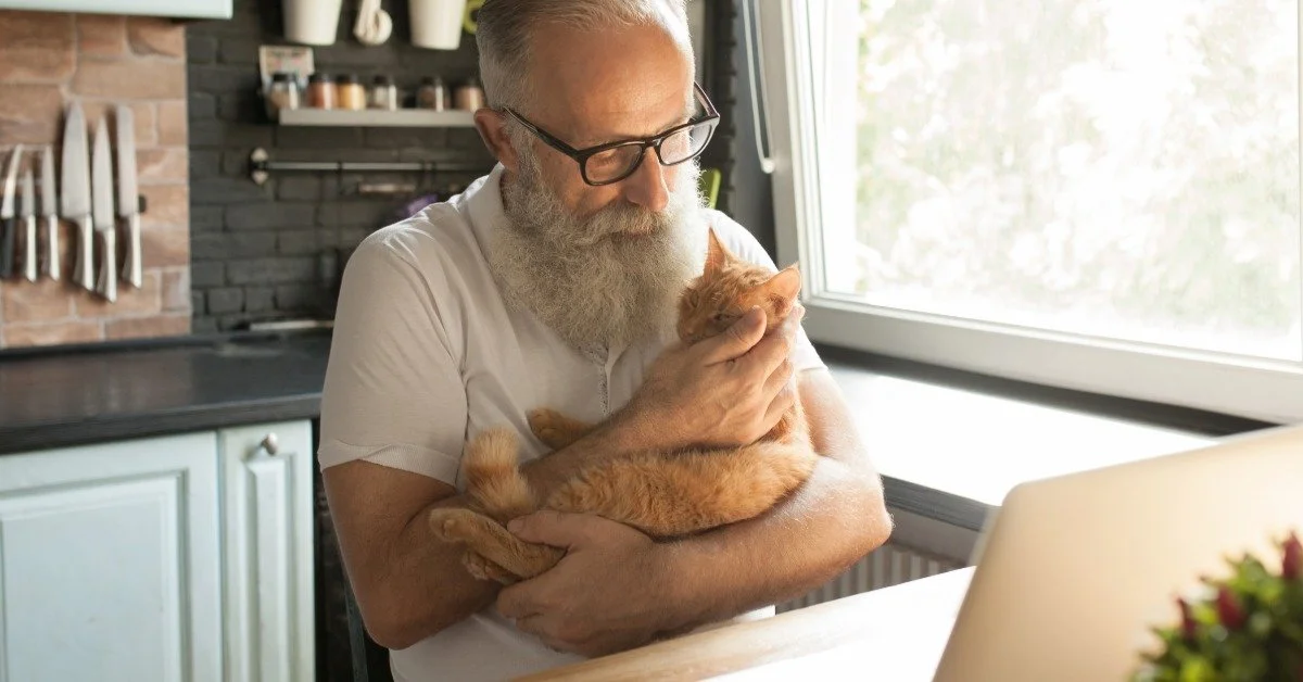 An older man with glasses holds a small orange cat while sitting at a kitchen table beside an open laptop near a window.