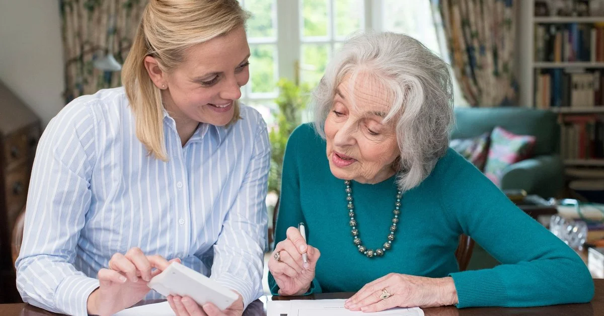 An elderly woman wearing a blue shirt holds a pen while looking at a piece of paper. A younger woman sits beside her.