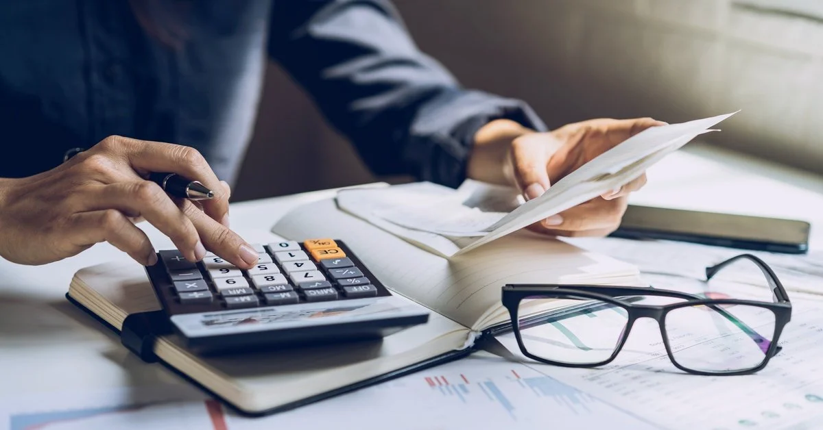 A person uses a calculator to review financial accounts at a desk, with a notebook, glasses, and documents spread out nearby.