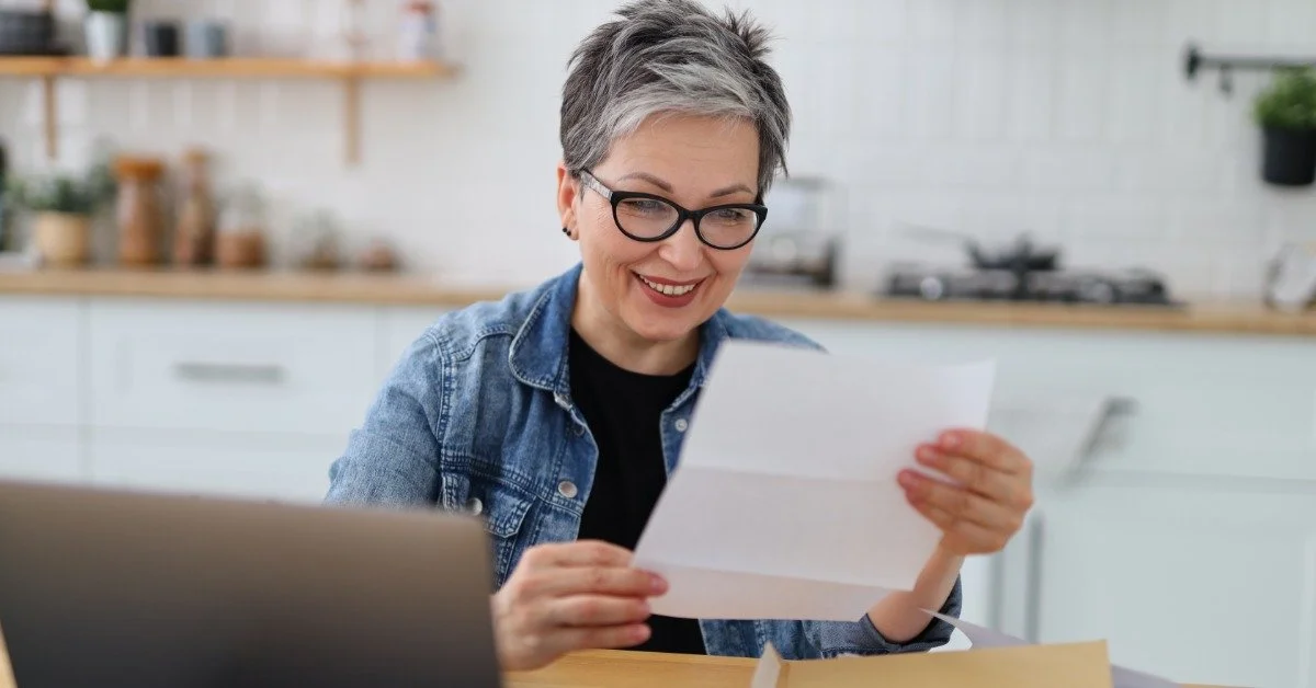 A smiling mature woman reviews a paper at a kitchen table while a laptop and folder sit open beside her.