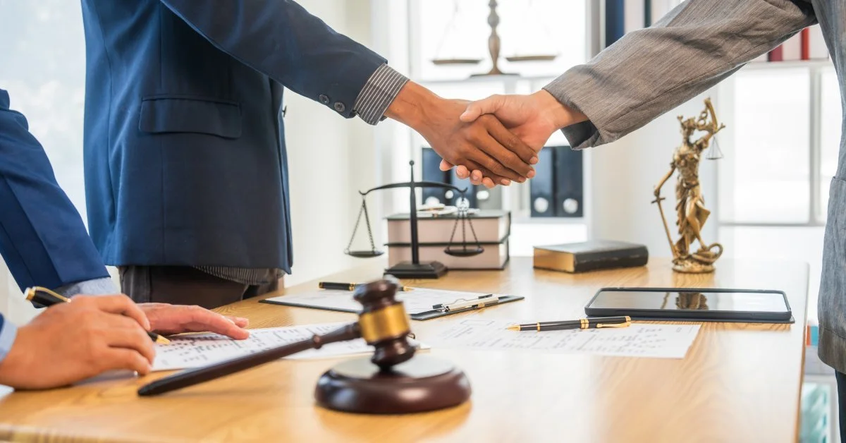 A probate lawyer shakes hands with their client over a wooden table with a gavel, the scales of justice, and documents on it.