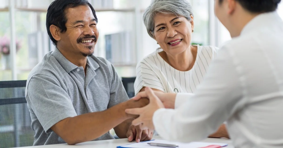 A smiling older couple shakes hands with a legal professional across a bright office table with documents nearby.