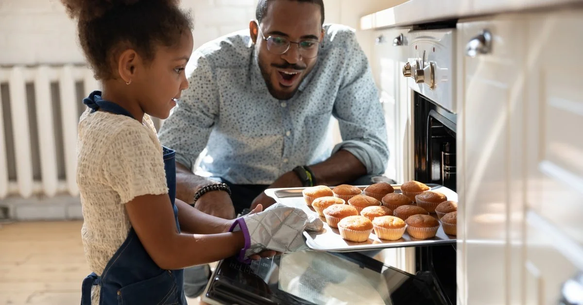 A man smiles in surprise as he watches a little girl take muffins out of the oven; she is wearing a little apron and an oven mitt.