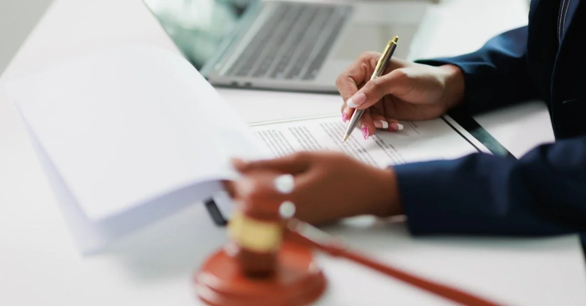 A person holding a pen over printed documents at a desk with an open laptop and a wooden gavel in front.