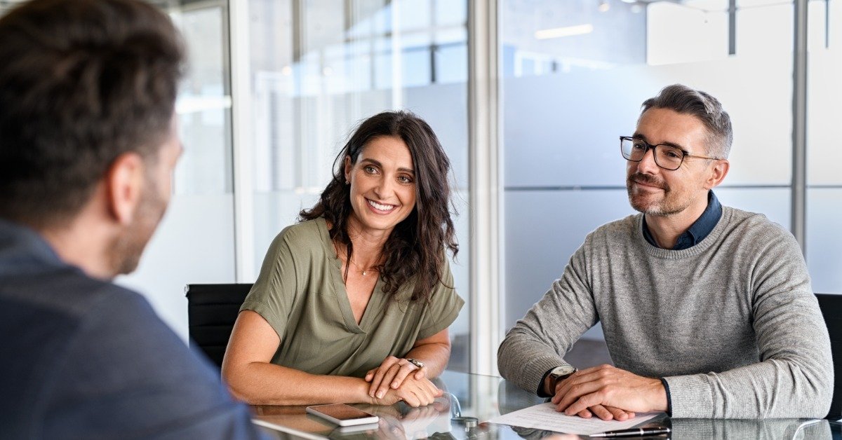 A mature couple smiles at an estate planning lawyer while reviewing documents across a glass table in a bright office.