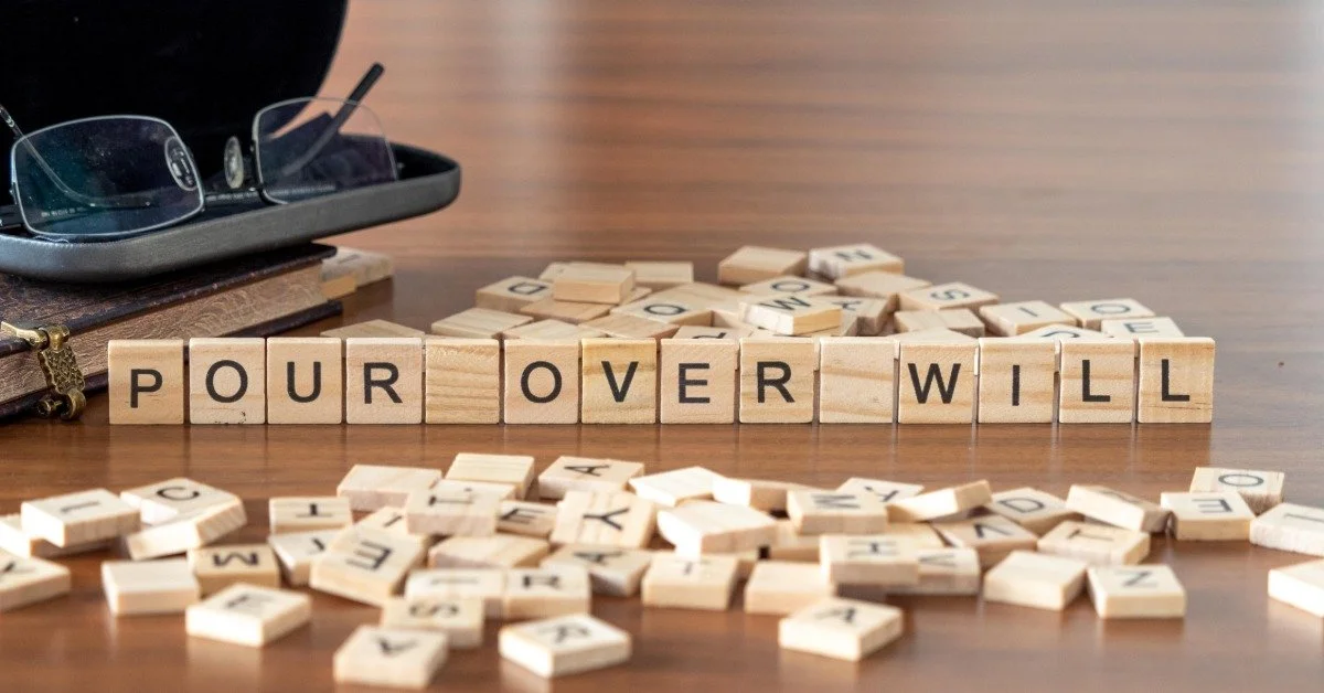 Wooden letter tiles spell the phrase "POUR OVER WILL" on a desk beside a glasses case and scattered letter tiles.