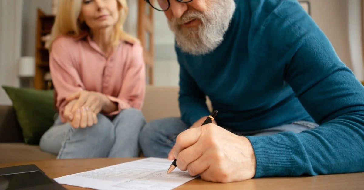 An older man signs a document at a wooden table while his wife watches closely sitting on a couch behind him.