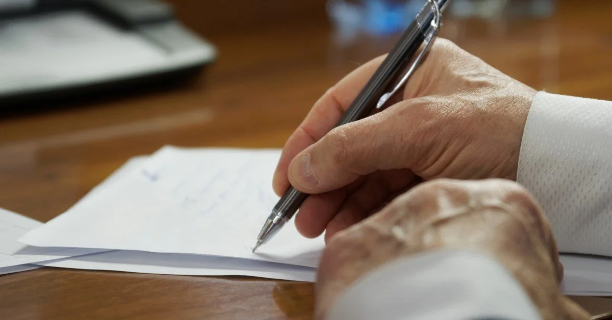 A close-up view of a hand signing legal documentation with a black pen on a light brown wooden desk.