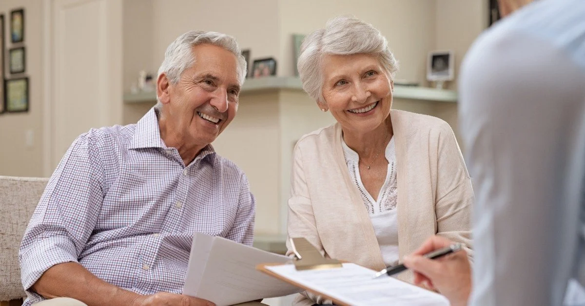 An older couple smiles as a financial advisor reviews documents with them in a bright, comfortable living room.
