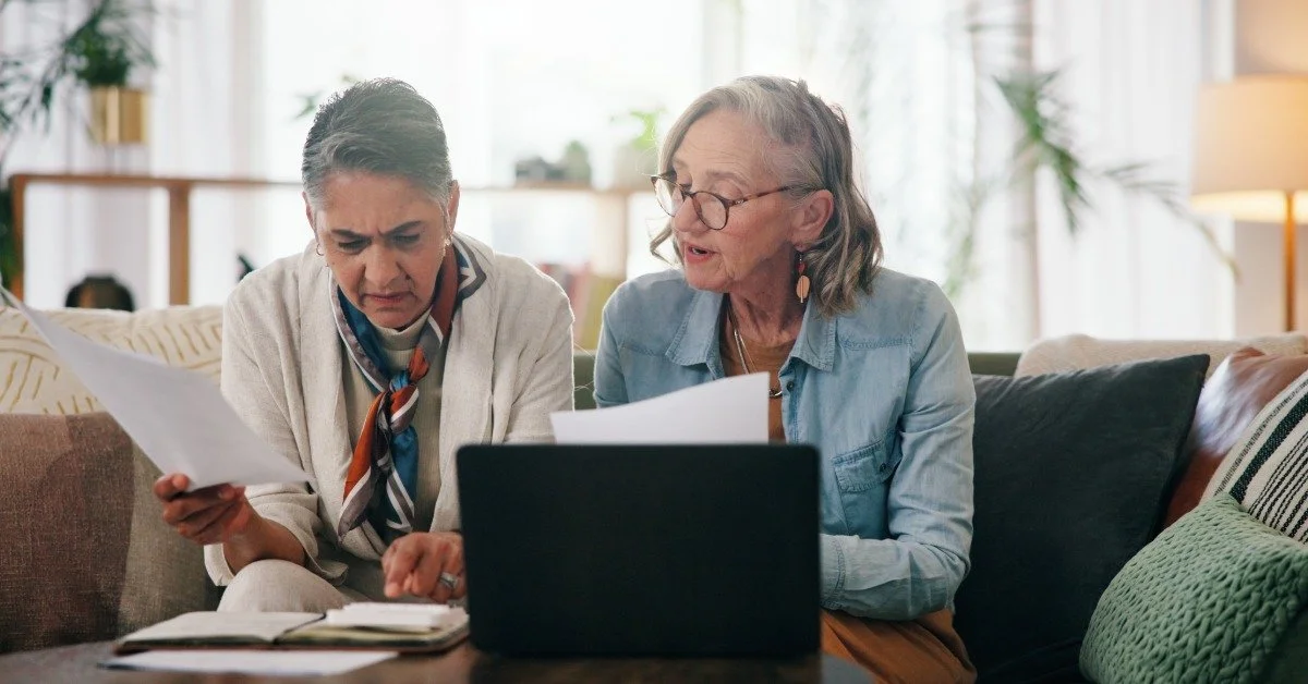 Two women review paperwork beside an open laptop while updating an estate plan in a bright living room.