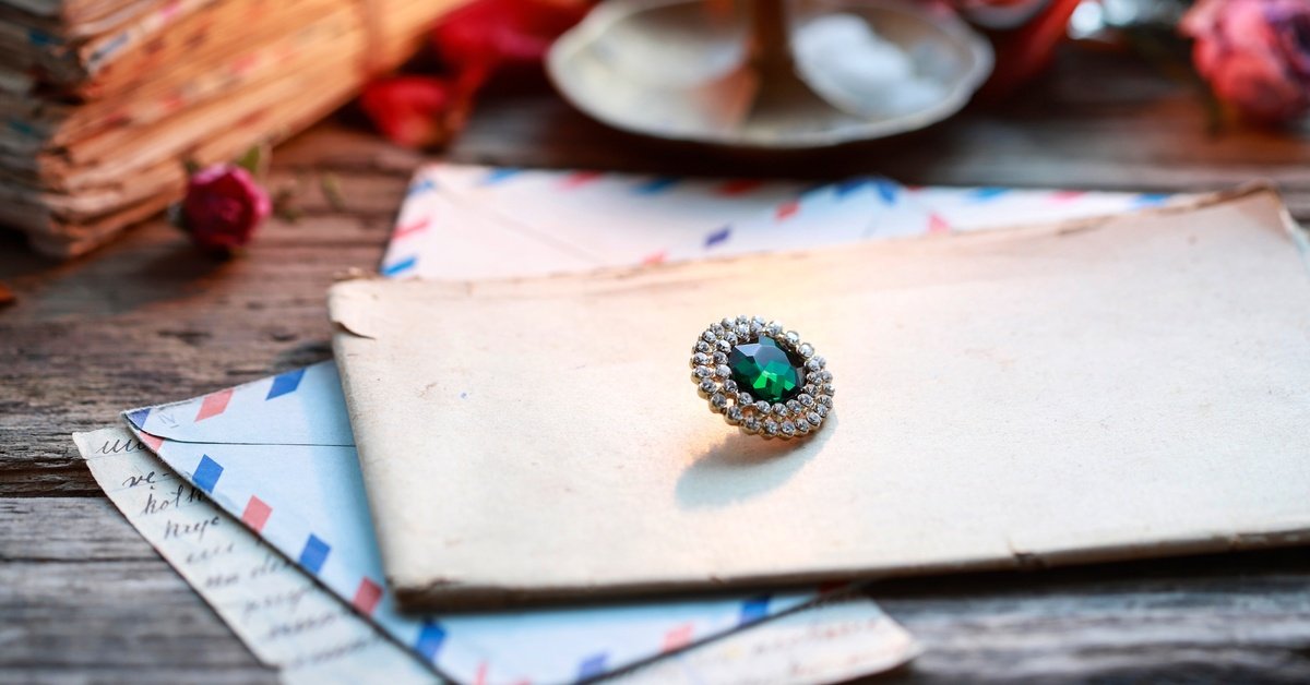 A vintage emerald brooch rests on old letters and envelopes atop a wooden table with red roses nearby.
