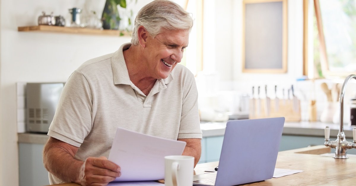 A smiling senior man reviews documents at a kitchen table with a laptop and coffee mug in a bright, modern kitchen.