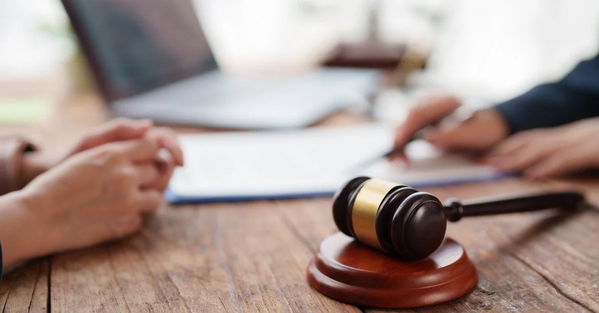 A wooden gavel rests on a table near papers and two people with hands visible during a document discussion.