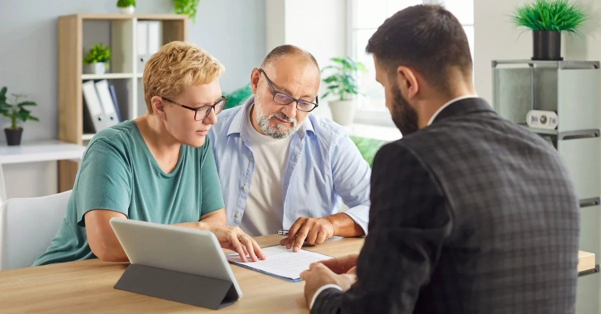 An older couple sits with a suited man at a wooden desk, looking over documents beside a tablet in a bright office.