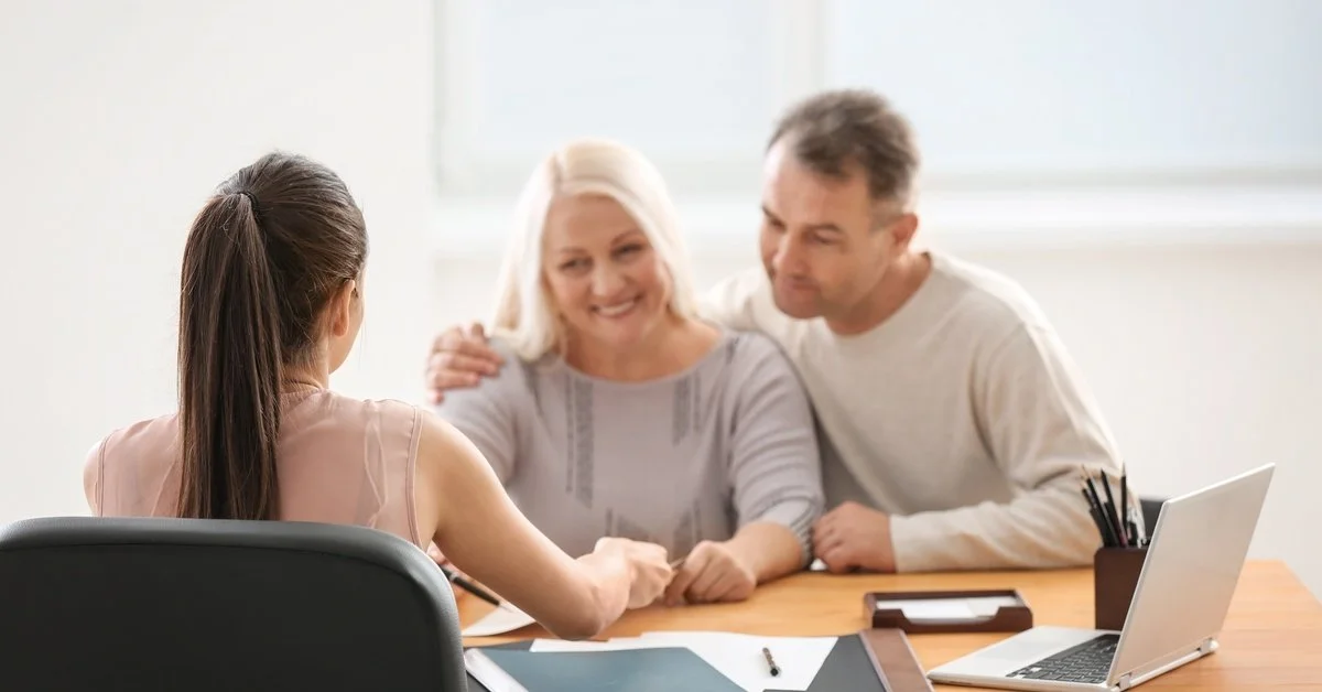 An older couple sit close to one another across from a younger woman. The table is covered in papers and a laptop.