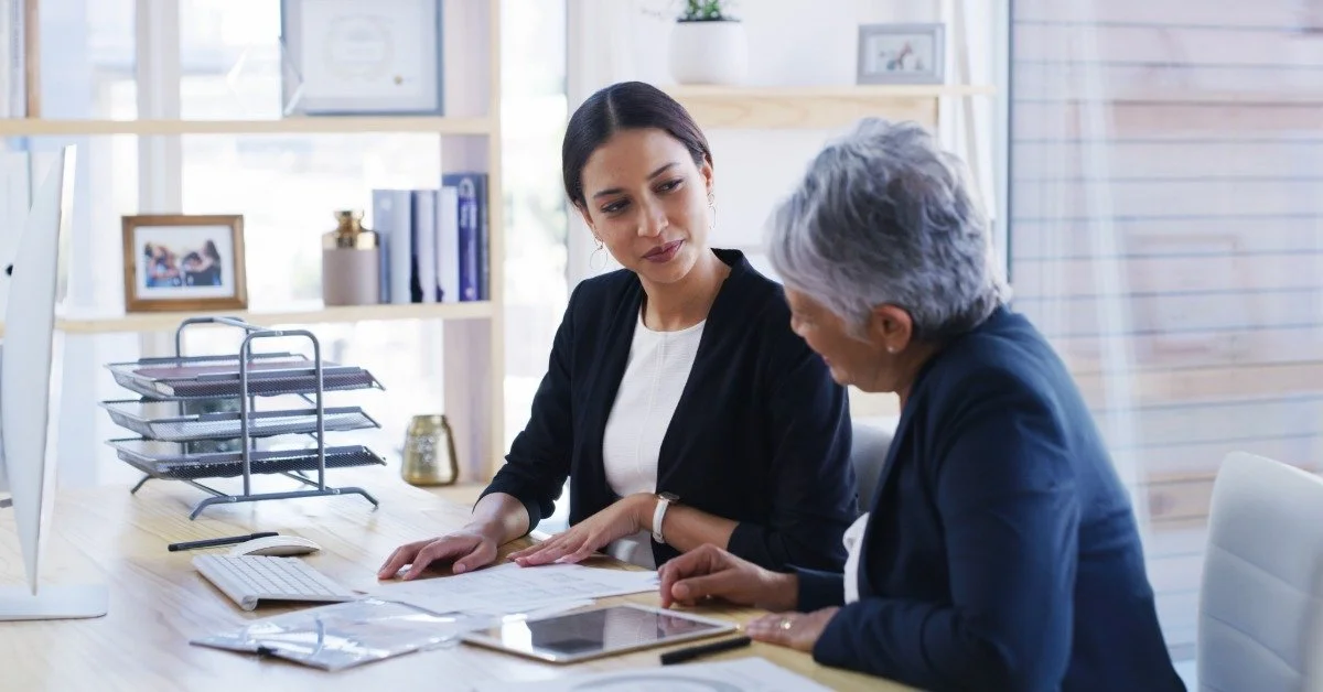 A female attorney reviews documents with an older woman as they sit together at a desk in a bright office.