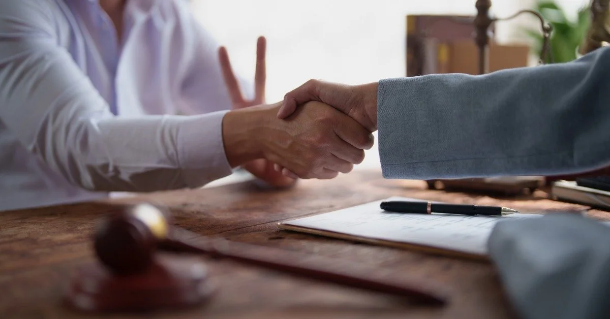 A client shakes a estate planning lawyer's hand across a legal desk with a gavel, a pen, and paperwork nearby.