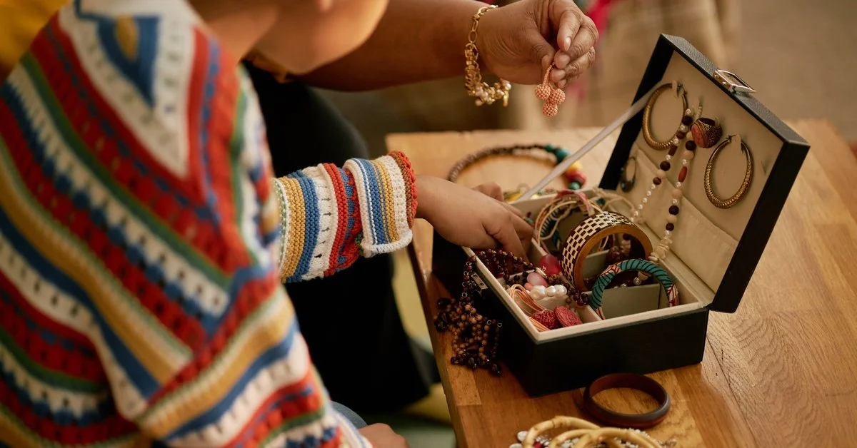 A woman and child sort through a jewelry box filled with bracelets, necklaces, and earrings on a wooden table.
