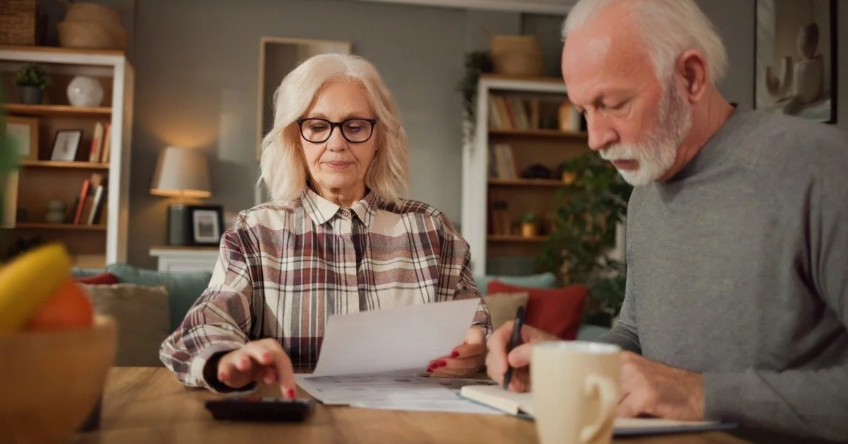 An older couple reviews a document and uses a calculator while planning finances together at a dining table.