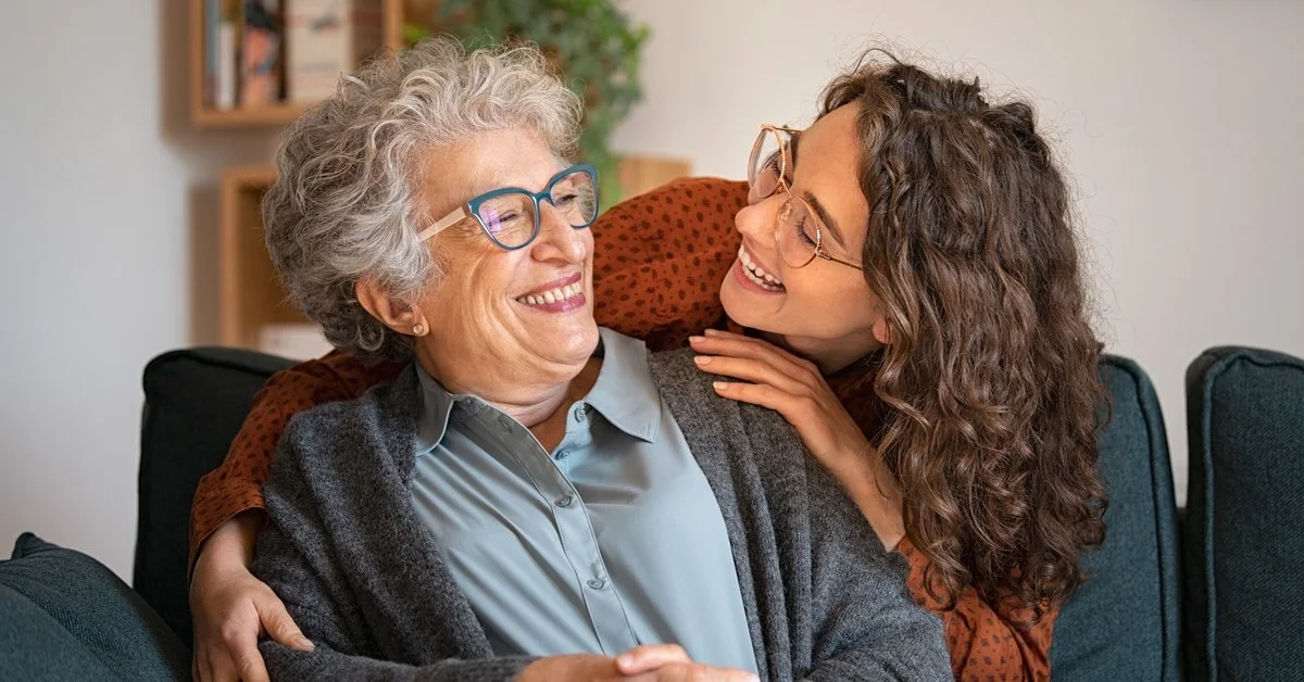 A smiling elderly woman sits in a chair, while a younger woman stands behind the chair hugging and looking at her.