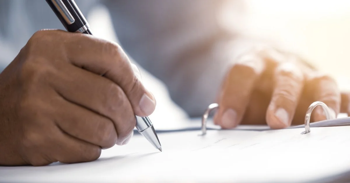 A close-up of a man's hand holding a silver pen. He is signing a piece of paper that's in a three-ring binder.