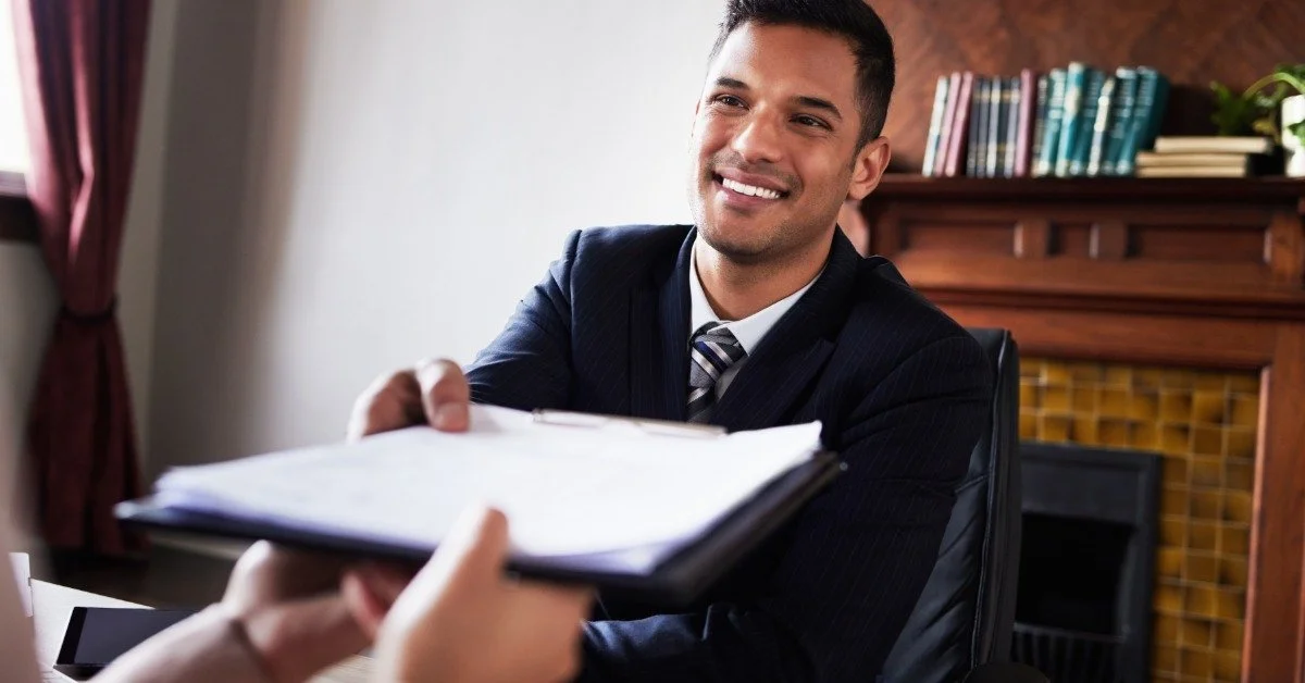 A male attorney hands a folder of documents to a client while smiling across a desk in a professional office.