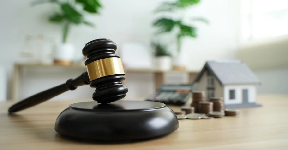 A judge's gavel rests on a light brown wooden desk with coins, a calculator, and a home blurred in the background.
