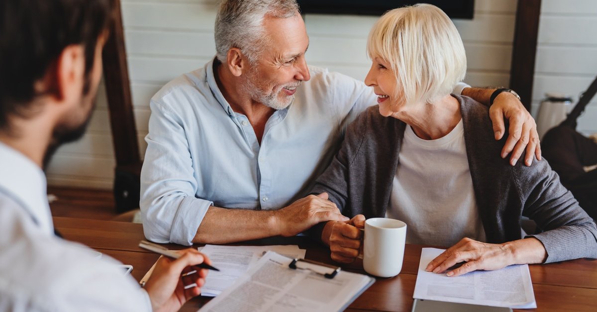 A lawyer consults an elderly couple at a wooden table with documents, a clipboard, and a tablet in an office.