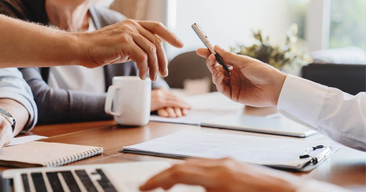 An older woman and an lawyer review estate documents at a desk as the lawyer holds a pen near a clipboard.