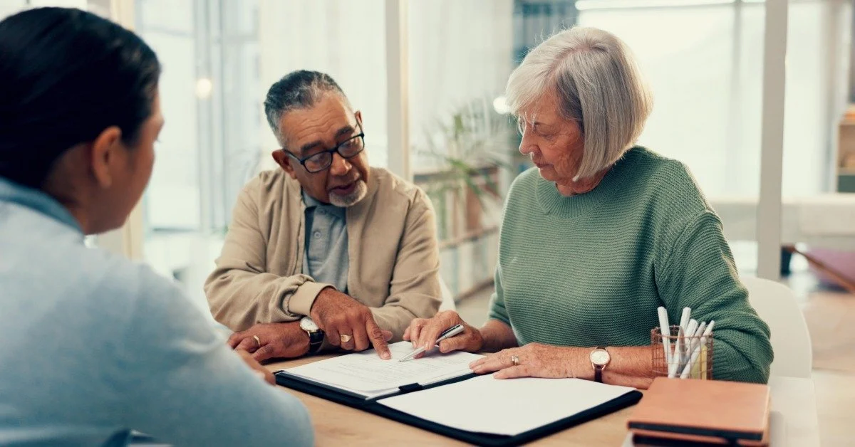 An older couple reviews and points to documents while an advisor guides them across a desk in a modern office.