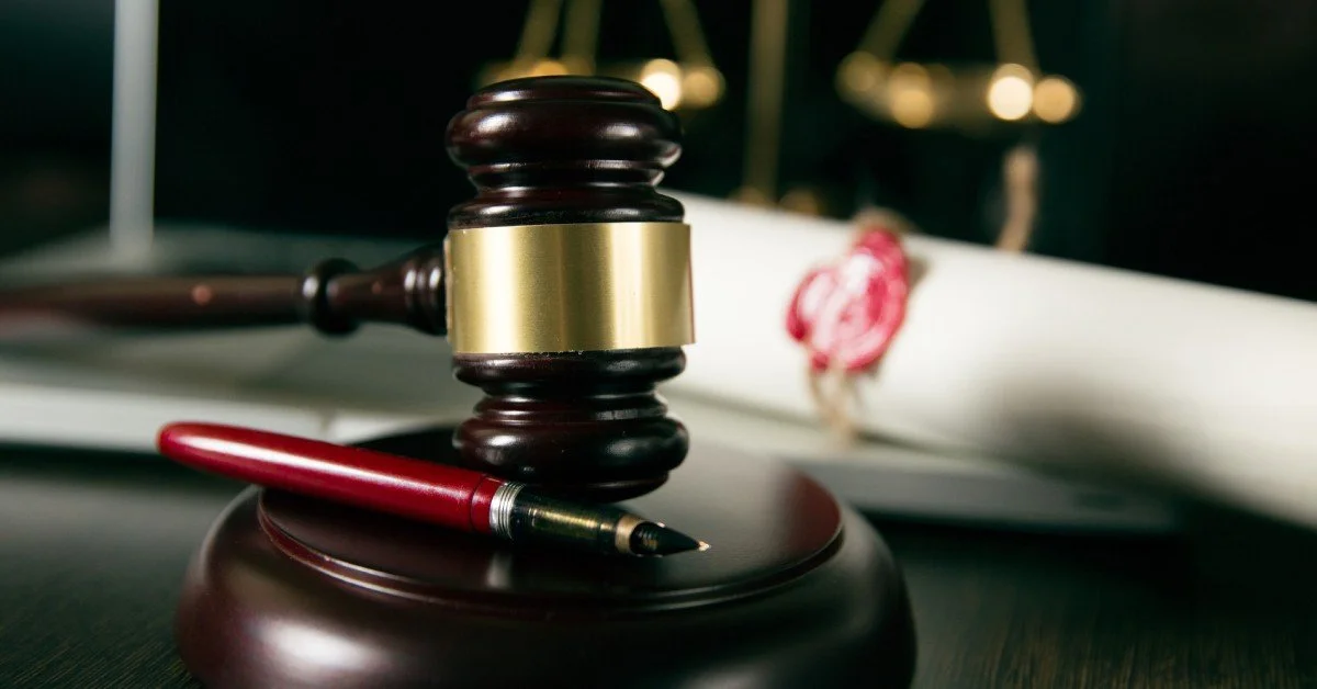 A wooden gavel and a red fountain pen rest on a dark surface, with blurred scales of justice in the background.