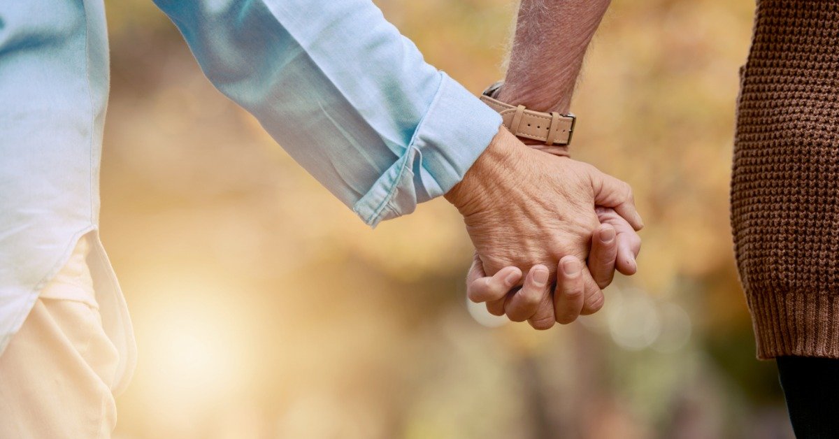 A close-up of an older couple holding hands while walking outdoors in a sunlit park with autumn trees in the background.