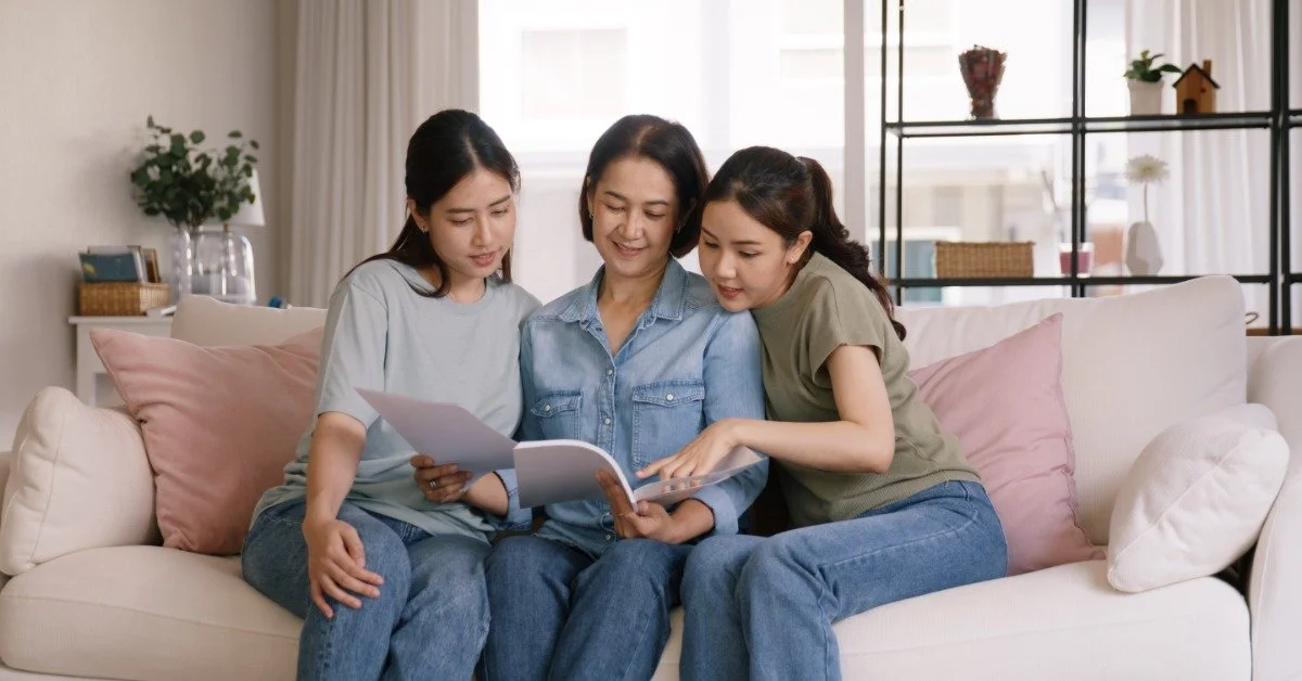 Three women sitting close together on a sofa while reading papers and a booklet, with pink cushions behind them.