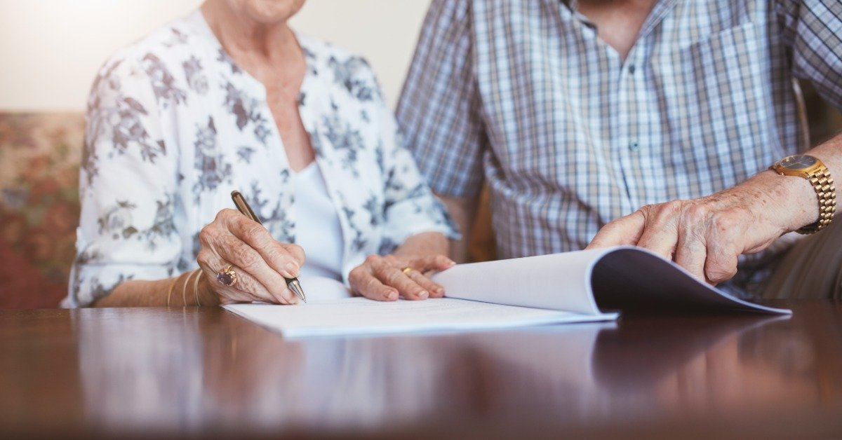 An older woman signs a document while a man turns the pages beside her at a table during an estate planning meeting.