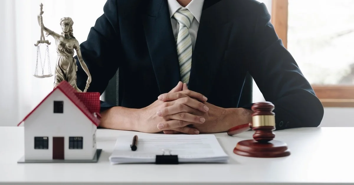 An estate lawyer sits at their desk with a gavel, paperwork, model house, and the scales of justice on it.