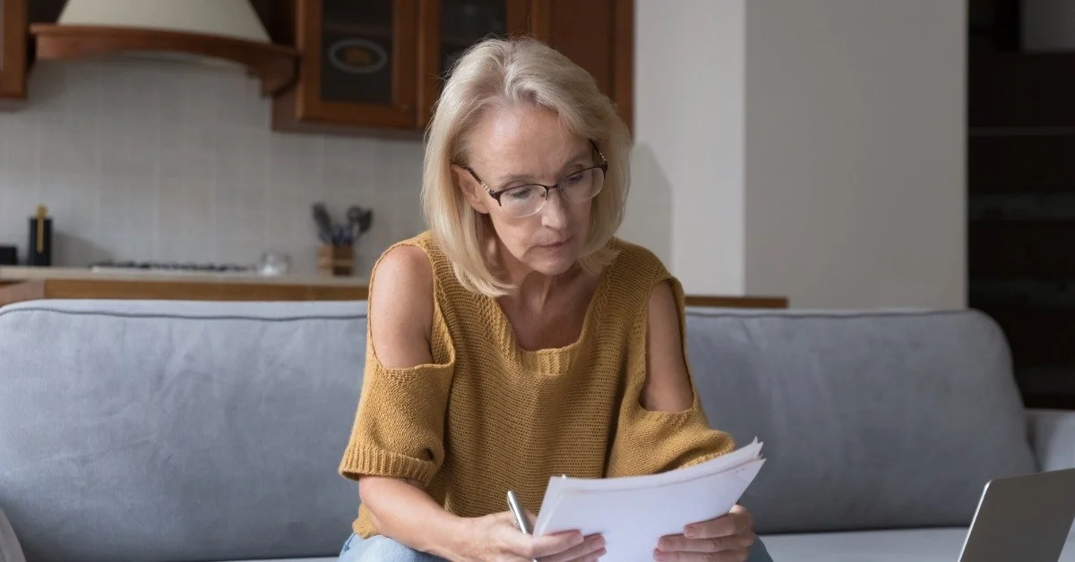 A senior woman wearing glasses reviews documents while sitting on a couch at home with a laptop nearby.