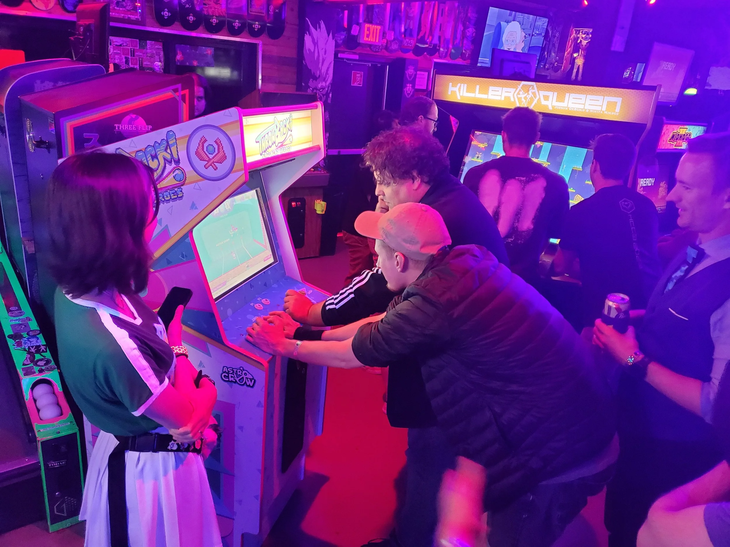 People playing classic arcade games in a neon-lit arcade with colorful lighting and screens, including "Throwback! Jai-Alai Heroes" and "Killer Queen."