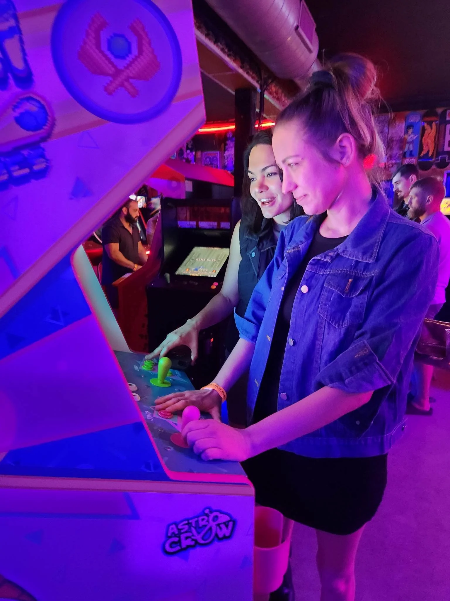 Two women play a retro arcade game "Throwback! Jai-Alai Heroes" together in a colorful, vibrant arcade setting with neon lighting.