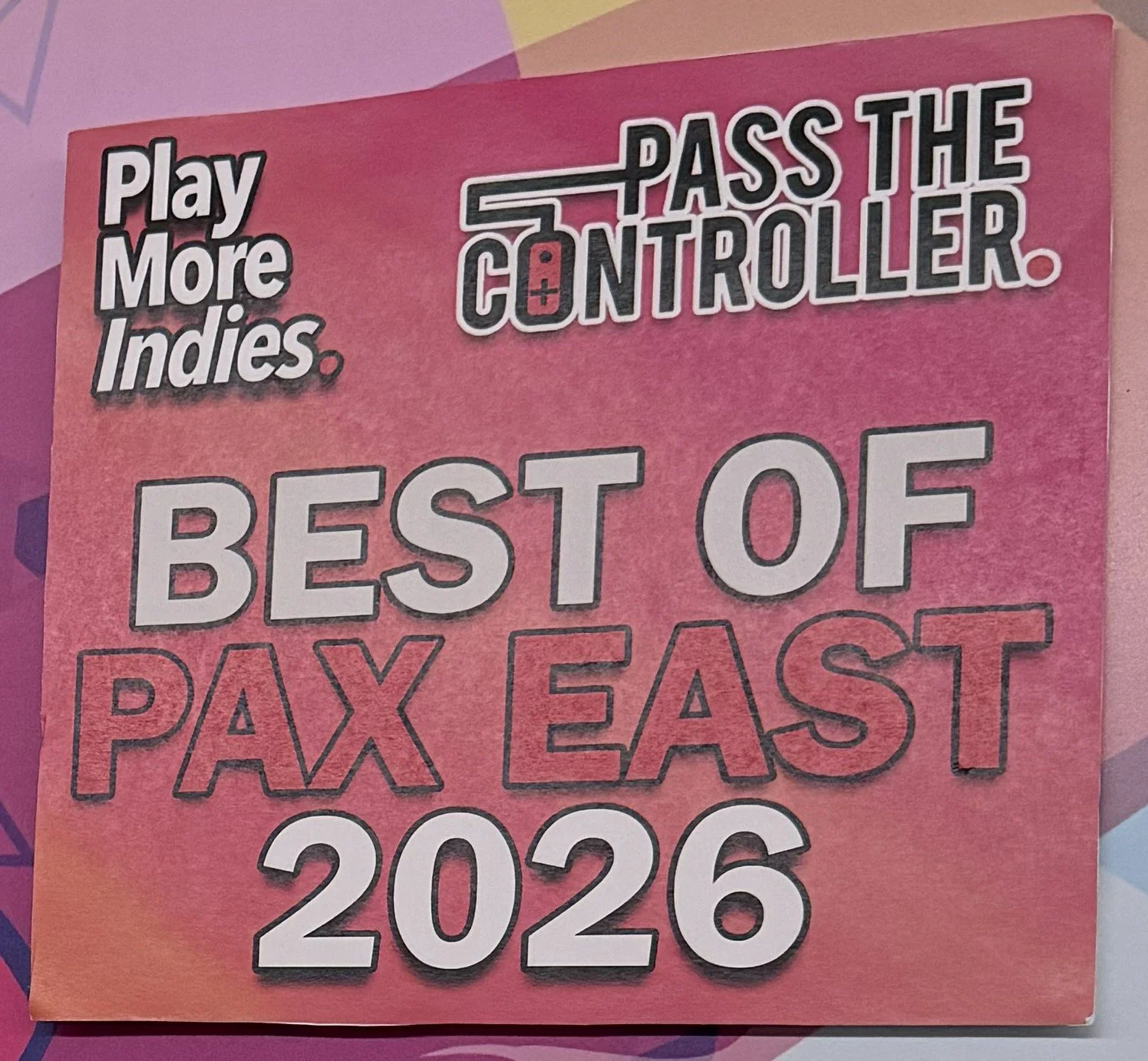 Pink promotional card with the message 'Play More Indies, Pass the Controller, Best of PAX East 2026' in various fonts and sizes.