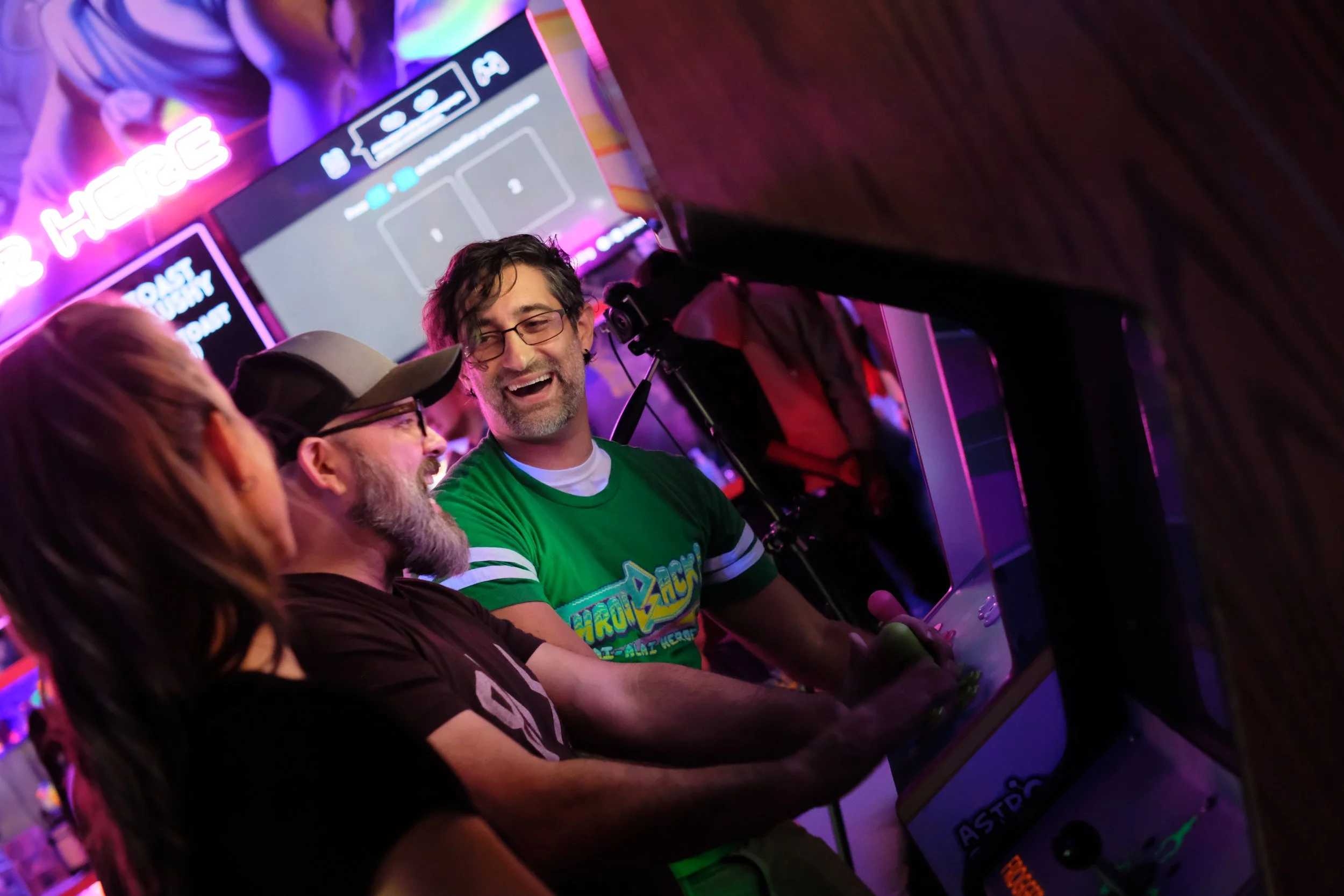 Three people sitting at a table, engaged in a conversation, with arcade game screens and neon signs in the background, illuminated by purple and pink lights.