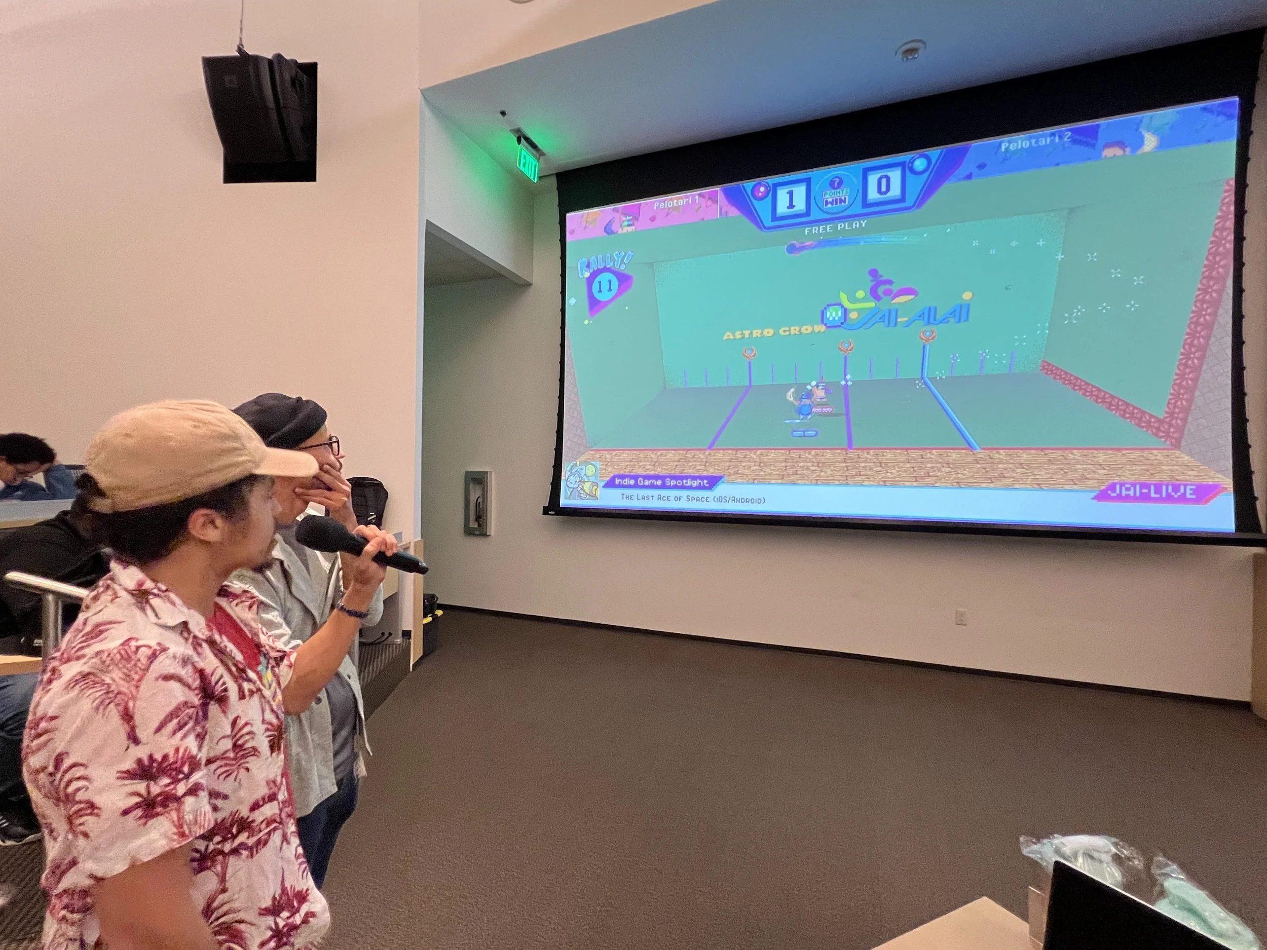 Two people are standing in front of a large screen displaying a colorful arcade-style game called "Throwback! Jai-Alai Heroes." One person is holding a microphone, and the other is covering their mouth with their hand, watching the game.