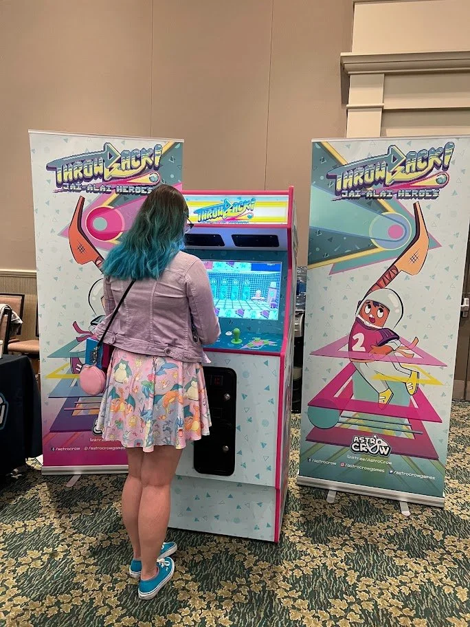A young woman with blue hair, wearing a pink jacket and a pastel skirt with unicorns, playing a retro arcade game called "Throwback! Jai-Alai Heroes" in a convention setting.