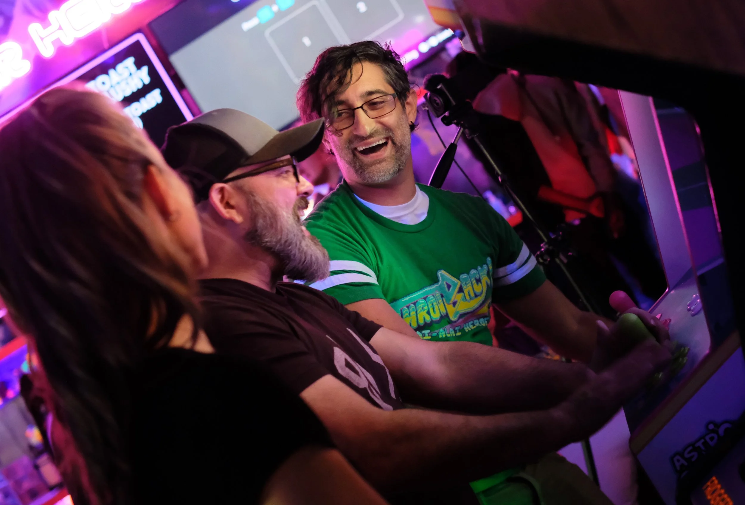 A group of people playing an arcade game at an arcade center with neon lights, including three men smiling and enjoying the game.