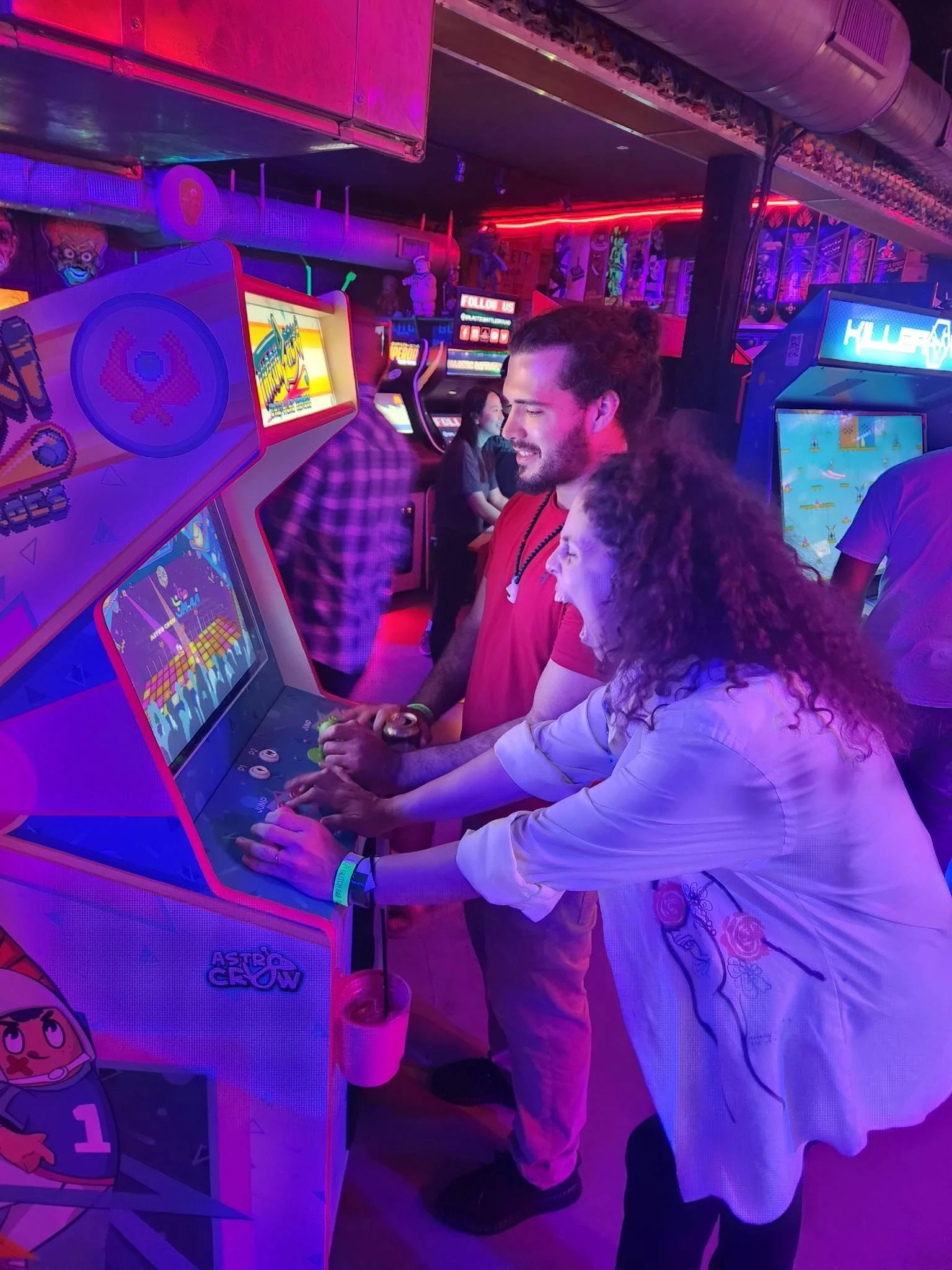 Two people playing a colorful arcade game "Throwback! Jai-Alai in a neon-lit arcade. The man has dark hair and a beard, wearing a red shirt, and the woman has curly hair, wearing a white top with floral embroidery, both intently focused on the game.