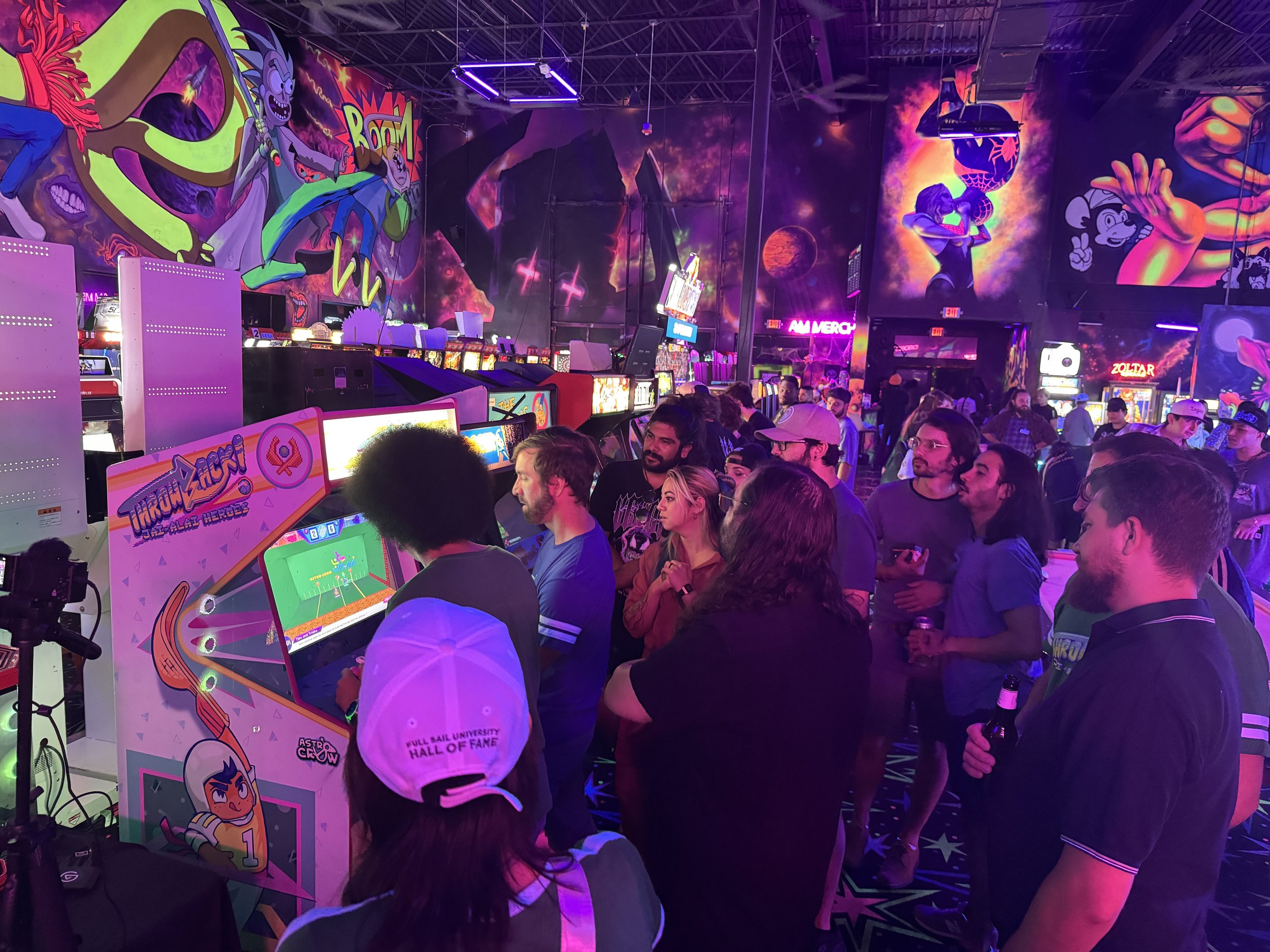 Inside an arcade with neon lighting, crowded with people playing arcade games, with colorful mural art on the walls featuring animated characters and space themes.