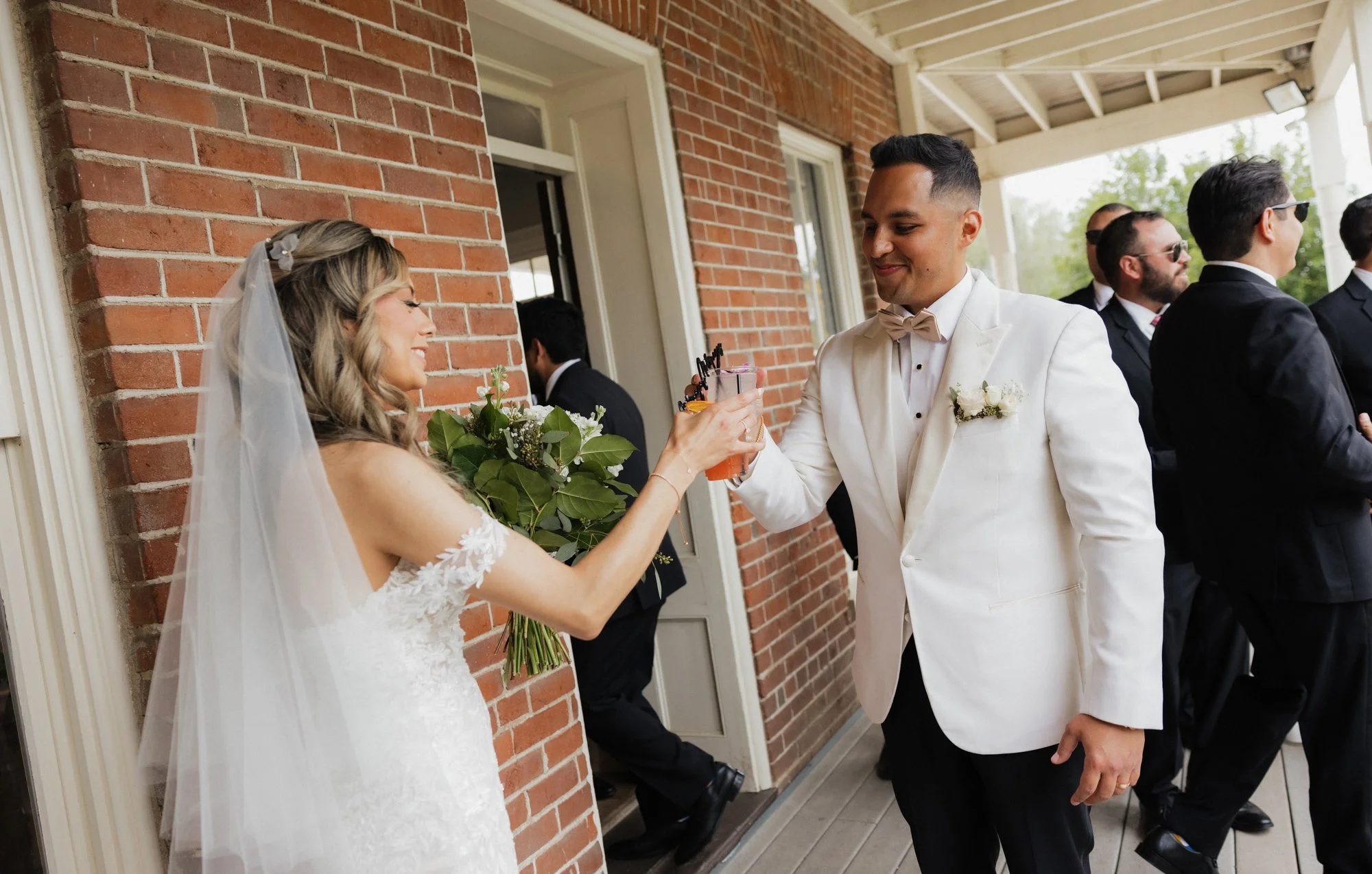 A bride holding a bouquet and a groom in a white tuxedo with a bow tie exchanging drinks during a wedding celebration outside on a covered porch with brick walls and several other people dressed in formal attire in the background at The Barton House.
