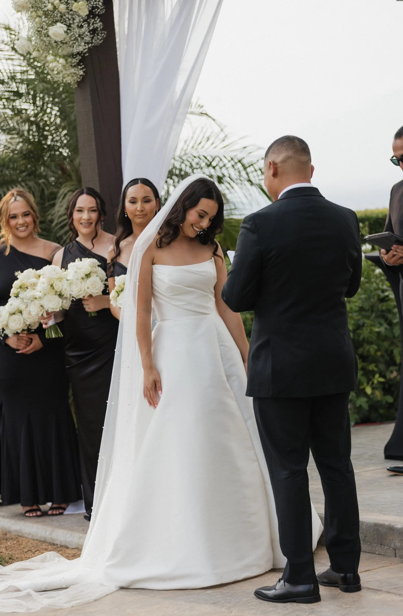 A bride and groom exchanging vows at their outdoor wedding ceremony, with bridesmaids holding bouquets of white roses in the background in Southern California.